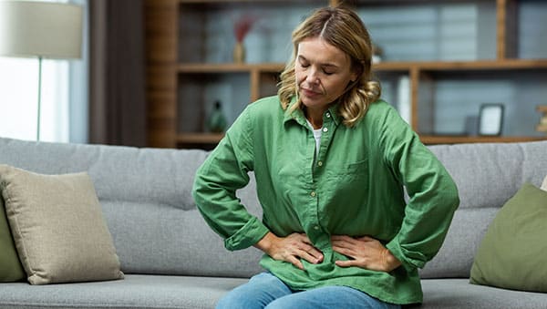 A woman is sitting on the sofa at home, holding her stomach with her hands in pain.
